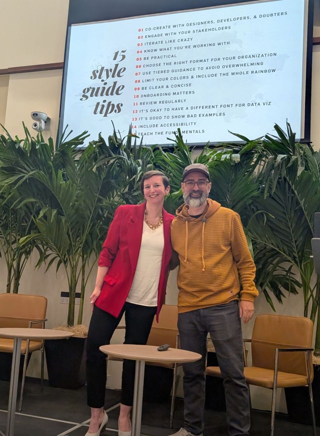 A picture of Alan and Amy at the Outlier conference in 2025 with a slide from the talk they gave with Max and Jon in the background.